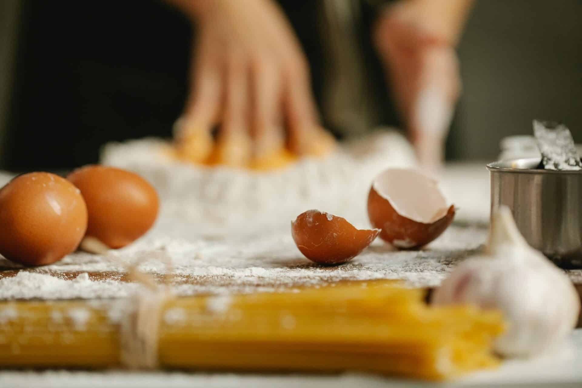 Ingredients Recipe Baked Egg Casserole Made with Crackers, including eggs, cracked eggshells, garlic, and flour, with hands preparing dough in the background.