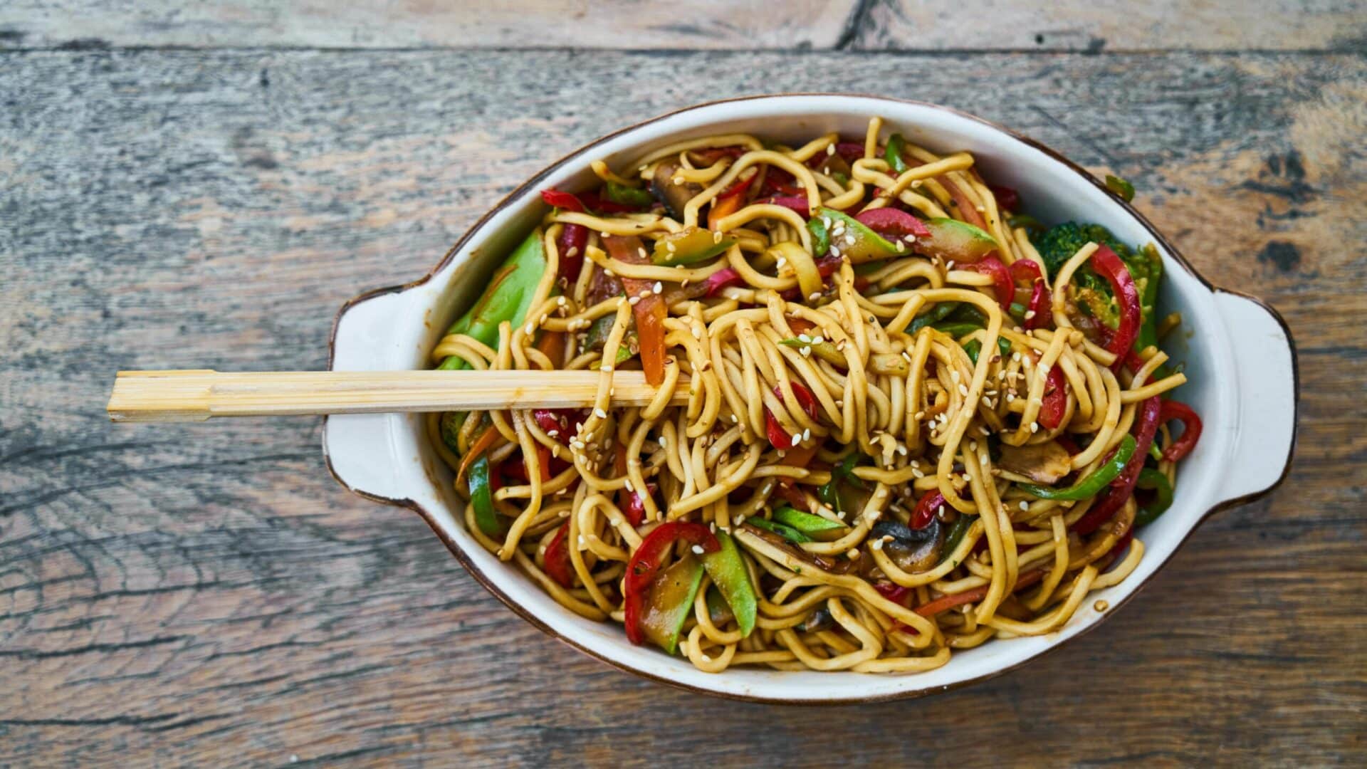 A bowl of Cup Noodles Stir Fry with fresh vegetables, sesame seeds, and chopsticks on a rustic wooden table.