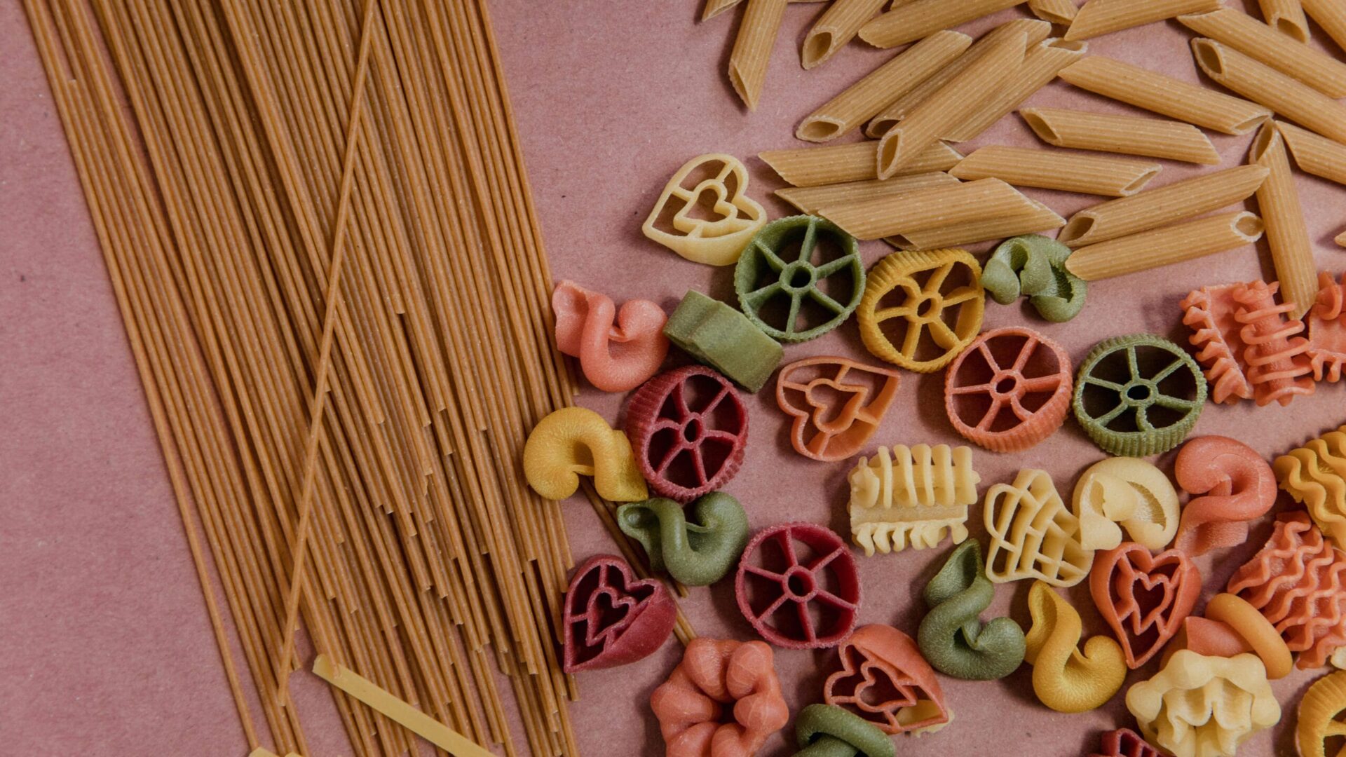An assortment of colorful heart shaped pasta alongside other pasta shapes, including penne and spaghetti, laid out on a textured surface.