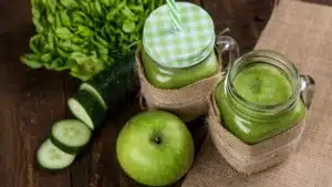Home 7 Fresh green detox smoothies in mason jars, wrapped in burlap, surrounded by cucumber, green apple, and lettuce on a rustic wooden background.
