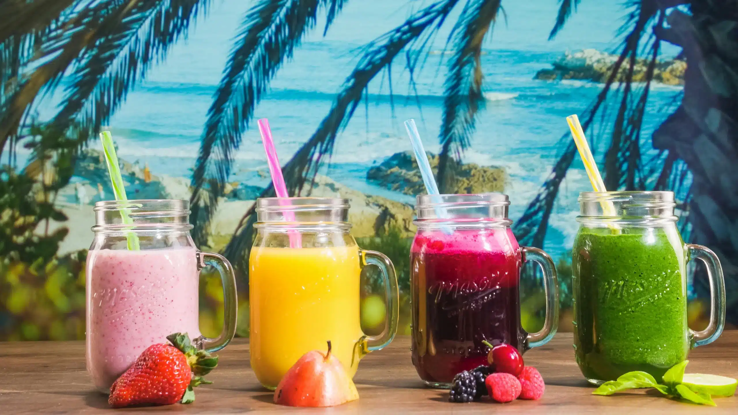 The Best Tropical Smoothie Recipes for a Refreshing Boost 9 Four colorful tropical smoothies in mason jars with straws, placed on a wooden table, framed by a picturesque beach and palm tree background.